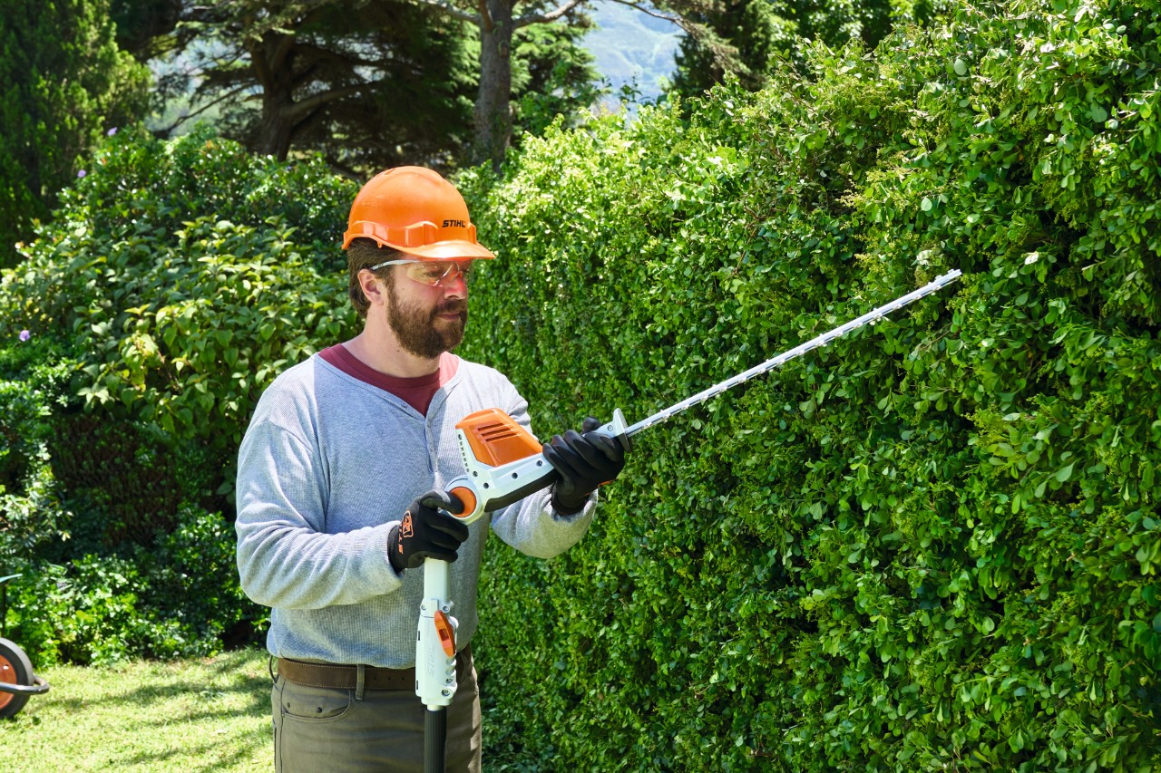 Un hombre con equipamiento de protección corta un seto alto con un cortasetos de altura de batería STIHL HLA 56