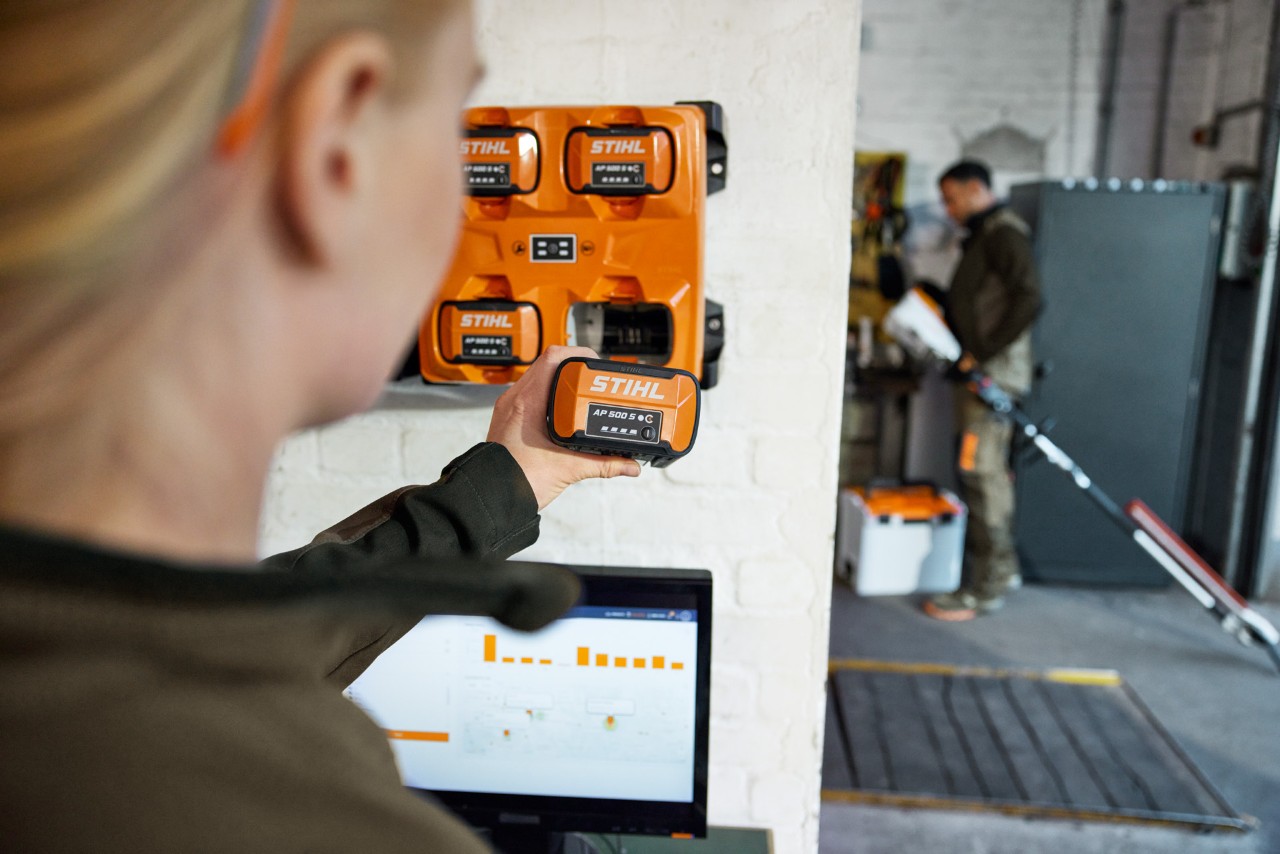Una mujer, delante de un ordenador portátil, toma una AP 300 S del cargador múltiple STIHL AL 301-4