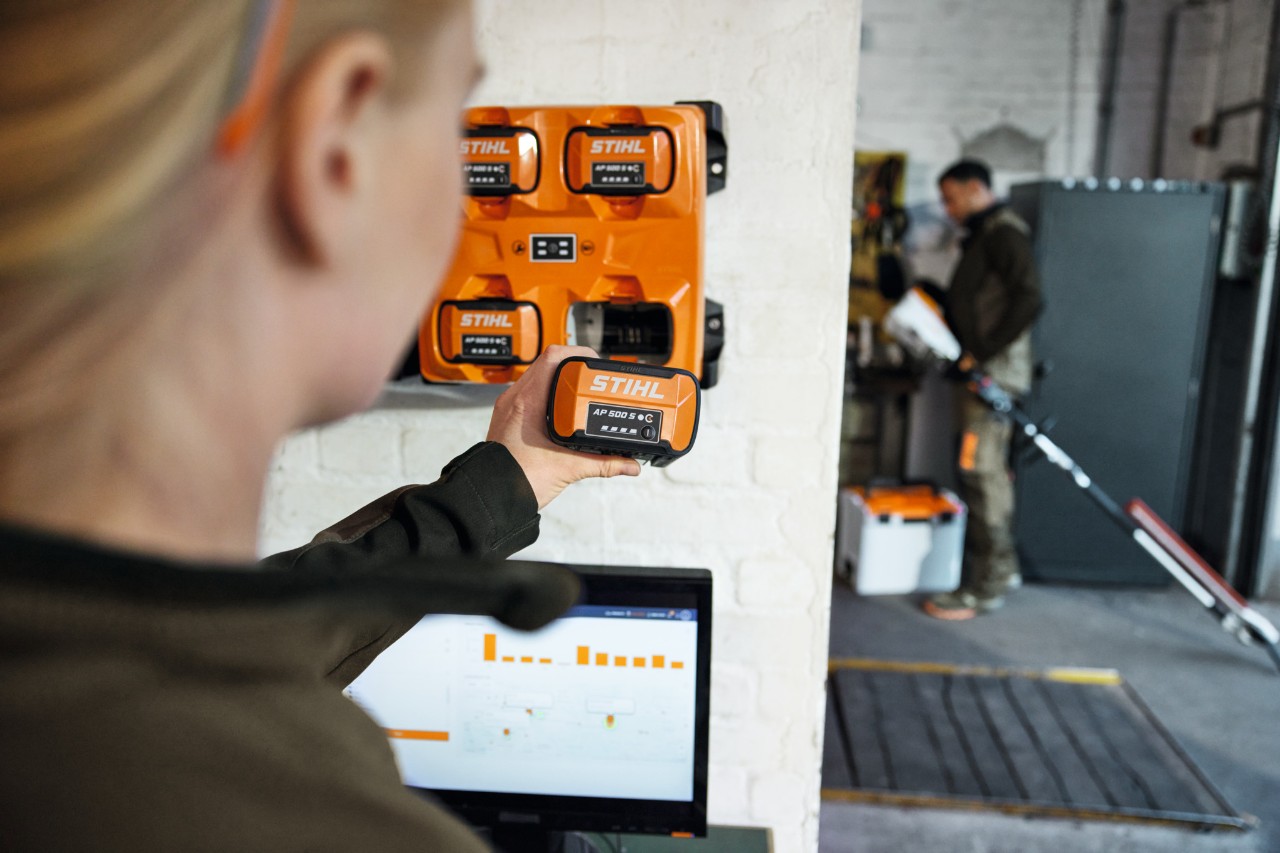 Una mujer, delante de un ordenador portátil, toma una AP 300 S del cargador múltiple STIHL AL 301-4